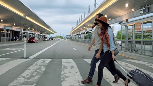 大笑的年轻时髦的夫妇走过人行横道到机场航站楼滚动行李箱。视频素材