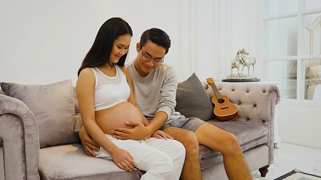 美丽的孕妇和她的丈夫抱着肚子在家里的沙发上听着宝宝，怀孕和家庭的概念视频下载