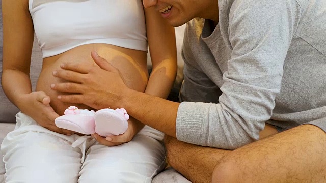 美丽的孕妇和她的丈夫抱着肚子在家里的沙发上听着宝宝，怀孕和家庭的概念视频下载