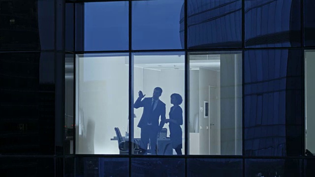 从摩天大楼外的航拍镜头:商人和女商人站在办公室窗户边谈生意。傍晚飞行拍摄的商务区和商务人士在大城市工作。视频素材
