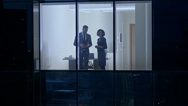 从摩天大楼外的航拍镜头:商人和女商人站在办公室窗户里交谈。找回傍晚商业区的飞行镜头和在大城市工作的商人。视频素材