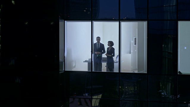 从摩天大楼外的航拍镜头:商人和女商人站在办公室窗户里交谈。飞行拍摄的商务区在晚上和商务人士在大城市工作。视频素材