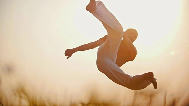 运动员跳着卡波耶拉舞，在美丽的夏日日落背景下表演一个困难的跳跃视频下载