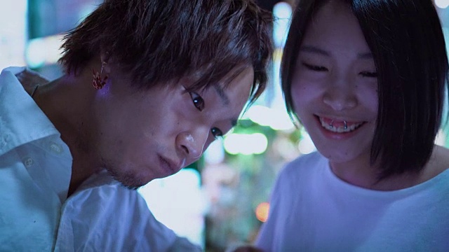 日本一对年轻的男孩和女孩聊天，大笑，使用手机和共享屏幕。在背景模糊的广告广告牌和城市灯光在晚上。视频素材