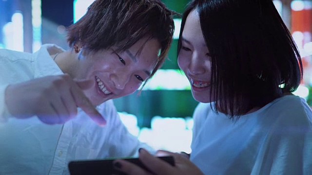 日本一对年轻的男孩和女孩聊天，大笑，使用手机和共享屏幕。在背景模糊的广告广告牌和城市灯光在晚上。视频素材