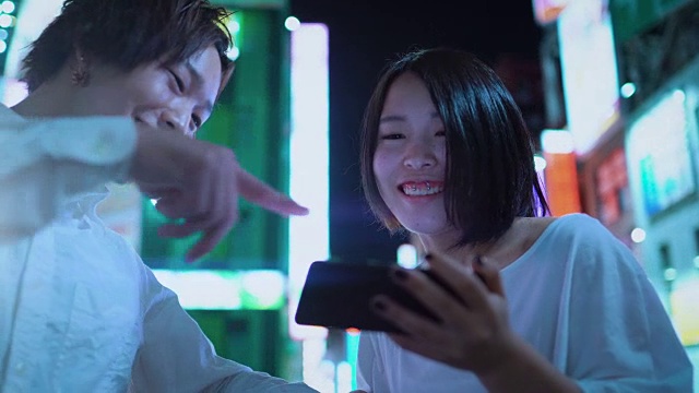 年轻的东亚男孩和女孩夫妇聊天，大笑，使用手机和共享屏幕。在背景模糊的广告广告牌和城市灯光在晚上。视频素材