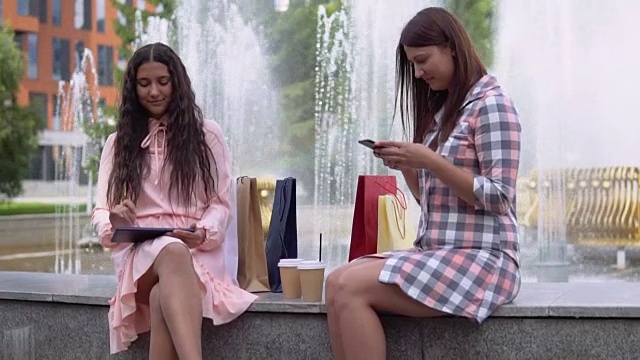 两个女孩购物后坐在公园的喷泉附近。缓慢的运动。高清视频素材