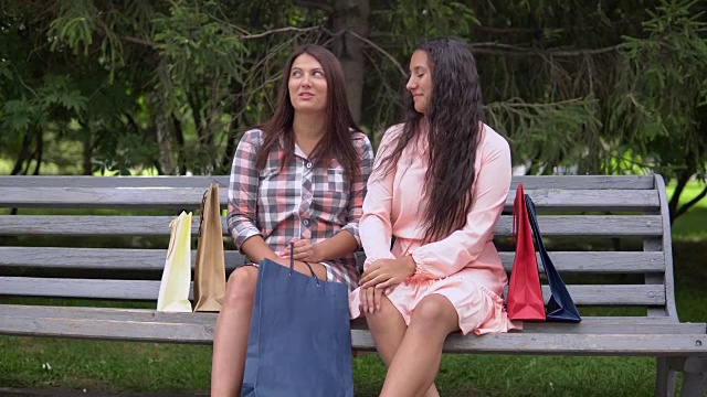 两个女孩购物后坐在公园的长椅上讨论买的东西。4 k视频素材