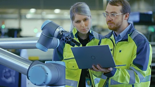 在工厂:女总生产工程师和男自动化工程师使用笔记本电脑为机器人手臂编程，并讨论努力的效率。自动化制造业的新时代。视频素材
