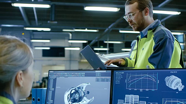 在工厂:男性和女性机械工程师在讨论机器人手臂制造项目时使用笔记本电脑和个人电脑。屏幕显示3D引擎概念与CAD软件。视频素材