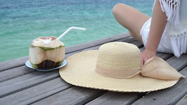女人戴着海帽她开心地坐着，在木桥上喝着椰子汁，看海边的沙滩，云和蓝天是无边无际的背景。视频下载