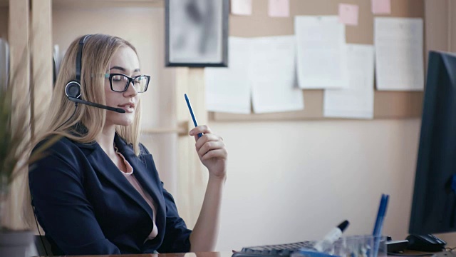 呼叫中心员工在办公室工作。一个金发姑娘戴着眼镜坐在办公室的桌子旁处理文件。女孩戴着耳机接电话，手里拿着一个自动手柄视频素材