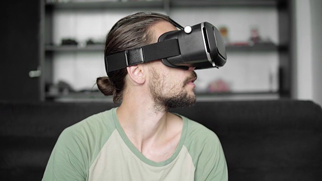 留着胡子的年轻潮男坐在客厅的沙发上，用他的VR头戴式显示器玩虚拟现实游戏或观看360视频。虚拟现实技术。视频素材