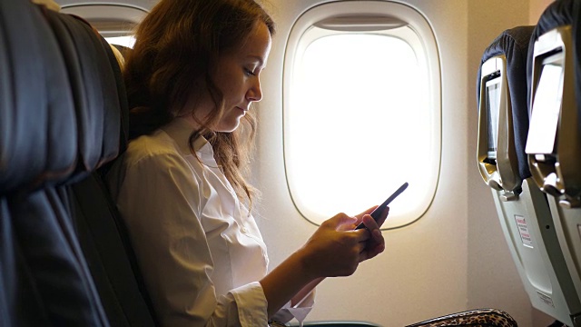 女游客在飞机上使用智能手机，坐在飞机靠窗的位置视频下载