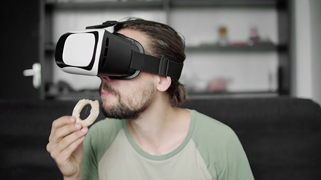 留着胡子的年轻潮男坐在客厅的沙发上吃着饼干，用VR头盔观看360度视频。虚拟现实技术。视频素材