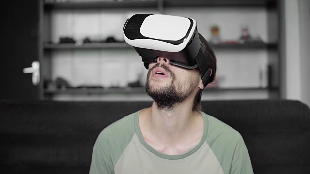 年轻的大胡子潮男使用他的VR头盔显示器进行虚拟现实游戏或观看360视频，并试图触摸他坐在沙发上看到的东西。虚拟现实技术。视频素材