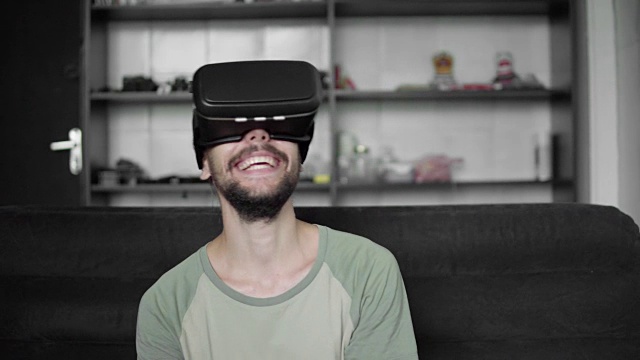年轻的大胡子潮男开始使用他的VR头盔显示器，戴着耳机玩虚拟现实游戏或观看360视频，坐在家里客厅的沙发上大笑。技术。视频素材