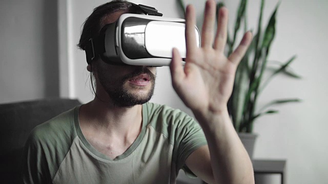 年轻的大胡子潮男使用他的VR头盔显示器进行虚拟现实游戏或观看360视频，并试图触摸他坐在沙发上看到的东西。虚拟现实技术。视频素材