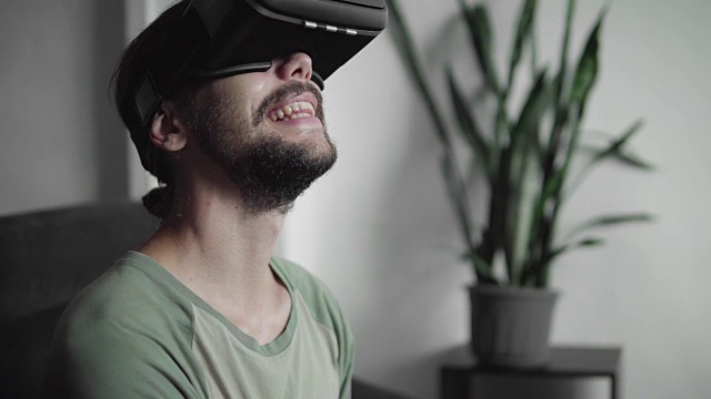 快乐的年轻大胡子潮人使用他的VR头盔显示虚拟现实游戏或观看360视频，而坐在家里的客厅沙发上。虚拟现实技术。视频素材