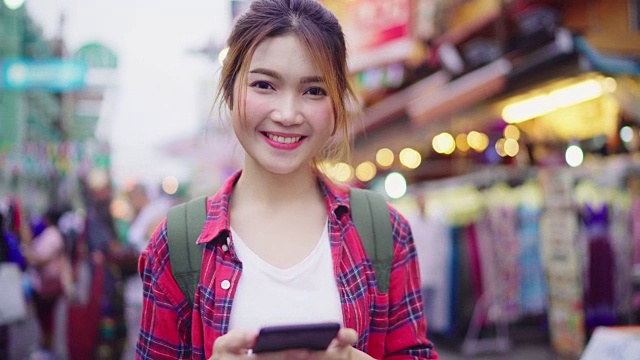 亚洲女性背包客微笑和使用智能手机独自旅行户外在曼谷考山路的城市街道，曼谷-泰国。视频下载