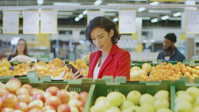 在超市:美丽的女人使用智能手机的肖像，在新鲜农产品通道中选择产品。女人沉浸在她的手机上网在背景彩色水果和有机蔬菜。视频素材