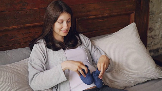 好玩的准妈妈在她怀孕的肚子上走着蓝色婴儿鞋，微笑着躺在家里阁楼风格的卧室的床上。怀孕和母性概念。视频下载