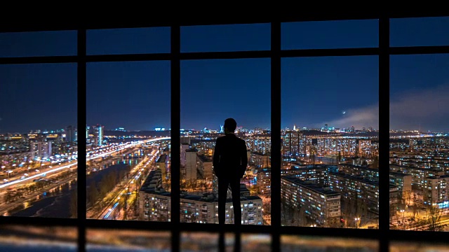 这个男人站在窗户旁边，背景是夜晚的大都市。时间流逝视频素材