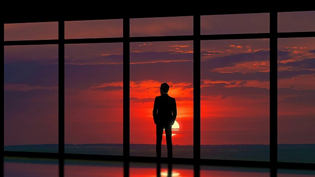 这个男人站在日落背景的全景窗户旁。时间流逝视频素材