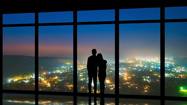 这对夫妻站在窗户旁，以夜色中的城市为背景。时间流逝视频素材