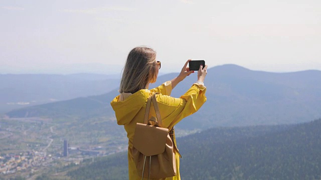 一名身穿黄色雨衣的女孩站在山顶用智能手机拍了一张照片，背景是蓝天和群山视频素材