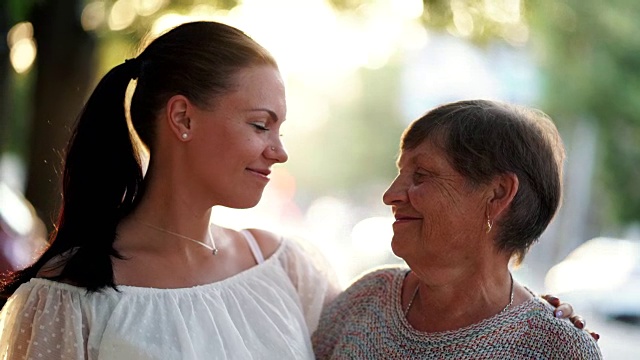 美丽的肖像祖母和她的孙女看着对方与爱在夏天的街道。太阳耀斑。女孩感激地拥抱奶奶。家庭的概念。4 k视频下载