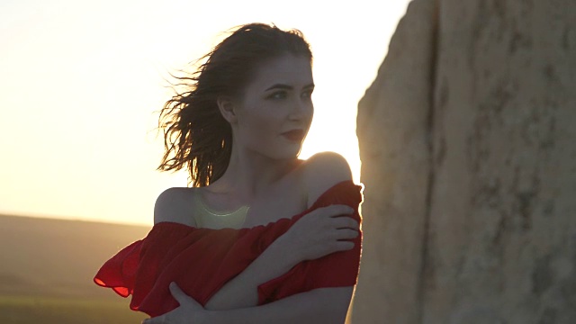 穿红衣服的年轻女子望着躲在悬崖后面的太阳，风吹散了她的头发视频素材
