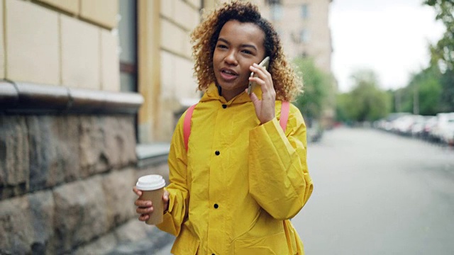 漂亮的非裔美国妇女正在喝着外带咖啡，用手机聊天，穿着鲜艳的夹克走在春天的城市里。现代科技和千禧一代的概念。视频下载