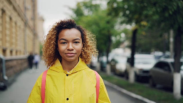 快乐的非洲裔美国女孩微笑的慢动作肖像，看着镜头站在户外绿树和美丽的建筑为背景。视频素材