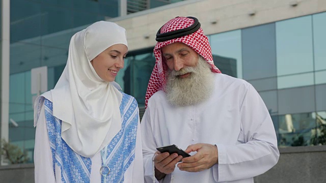 一个留着胡子的阿拉伯老年男子和一个年轻女孩在城市里用智能手机自拍视频下载