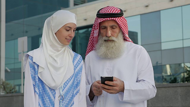 一个留着胡子的阿拉伯老年男子和一个年轻女孩在一个城市的智能手机上写一条信息视频下载