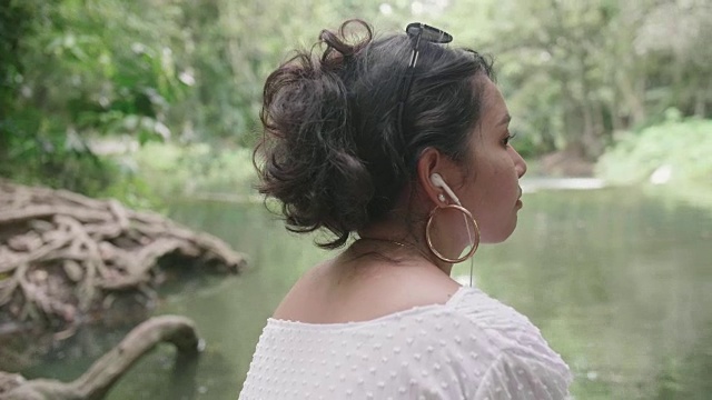 年轻女子坐在瀑布边，戴着耳机欣赏大自然。视频素材