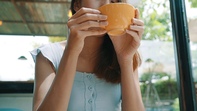 愉快的亚洲年轻女人喝温暖的咖啡或茶，坐在咖啡馆享受它。漂亮快乐的亚洲女人拿着一杯咖啡。视频素材