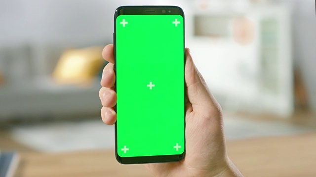 一个男人的手握绿色模拟屏幕智能手机的特写。现代移动电话。在舒适的客厅或家庭办公室的背景。视频素材