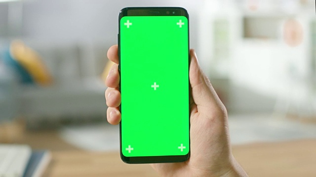 一个人拿着绿色模拟屏幕智能手机在他的手的特写。使用现代手机。在舒适的客厅或家庭办公室的背景。视频素材