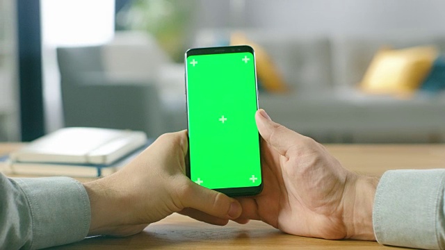 一个人拿着绿色模拟屏幕智能手机在他的手中的特写。使用现代高端设备。在舒适的客厅或家庭办公室的背景。视频素材