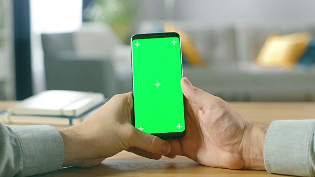一个人拿着绿色模拟屏幕智能手机在他的手中的特写。使用无线设备上网浏览。在舒适的客厅或家庭办公室的背景。视频素材