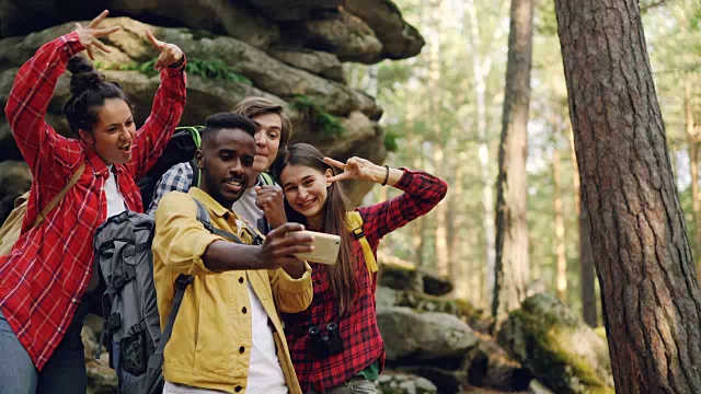 一名快乐的非裔美国人游客用智能手机和朋友们在大岩石附近的森林里自拍，人们做着滑稽的表情和手势。视频素材
