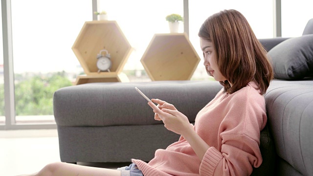 一名亚洲妇女躺在客厅的沙发上使用平板电脑。快乐的女性在家用平板电脑发短信、阅读、看视频和上网。生活方式女性在家的概念。视频素材