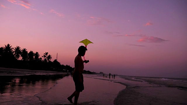 小男孩在日落时在海滩上玩风筝，慢镜头。视频素材