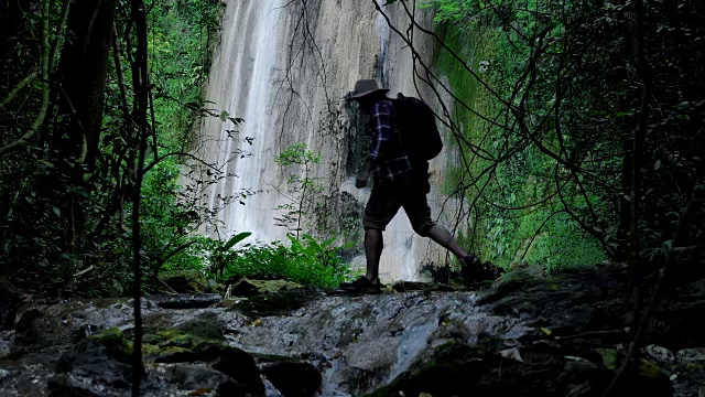 冒险家亚洲男性。在春天的森林里旅行，在阳光下发现瀑布。穿着舒适的衬衫，戴着帽子，背着背包，享受野外度假的生活方式。视频慢动作视频素材