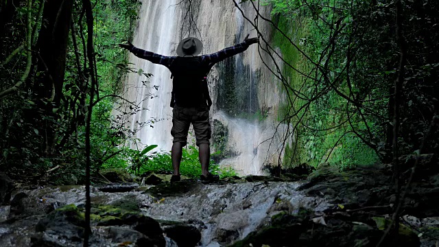 曼亚享受在瀑布前举起手旅行的生活方式和成功的概念度假到野外穿着舒适的衬衫，帽子与背包。视频慢动作视频素材