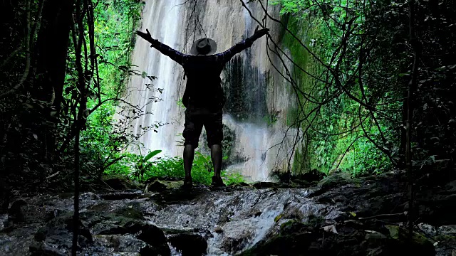 冒险家亚洲男性。在森林中旅行，阳光下的瀑布。旅行的生活方式和成功的概念度假到野外穿着舒适的衬衫，帽子和背包。视频素材