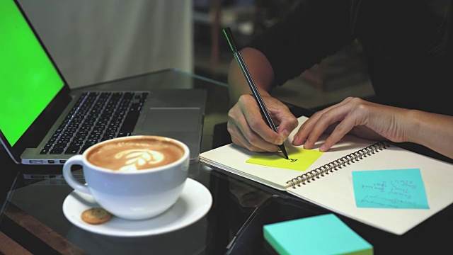 4k镜头镜头特写亚洲女商人的手工作和书写在便利贴笔记本电脑与绿屏笔记本电脑和咖啡杯在咖啡馆，生活方式和商业概念视频素材