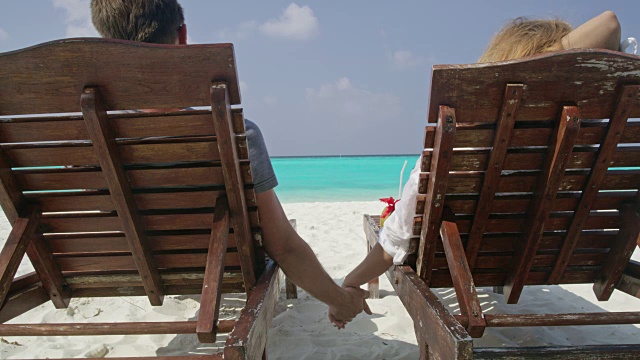 在马尔代夫阳光明媚的热带海滩上，一对夫妇手牵着手坐在躺椅上放松视频素材